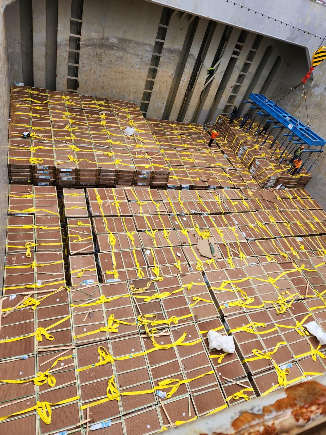 A ship's hold filled edge to edge with strapped plywood units, a spreader bar lifting a set out